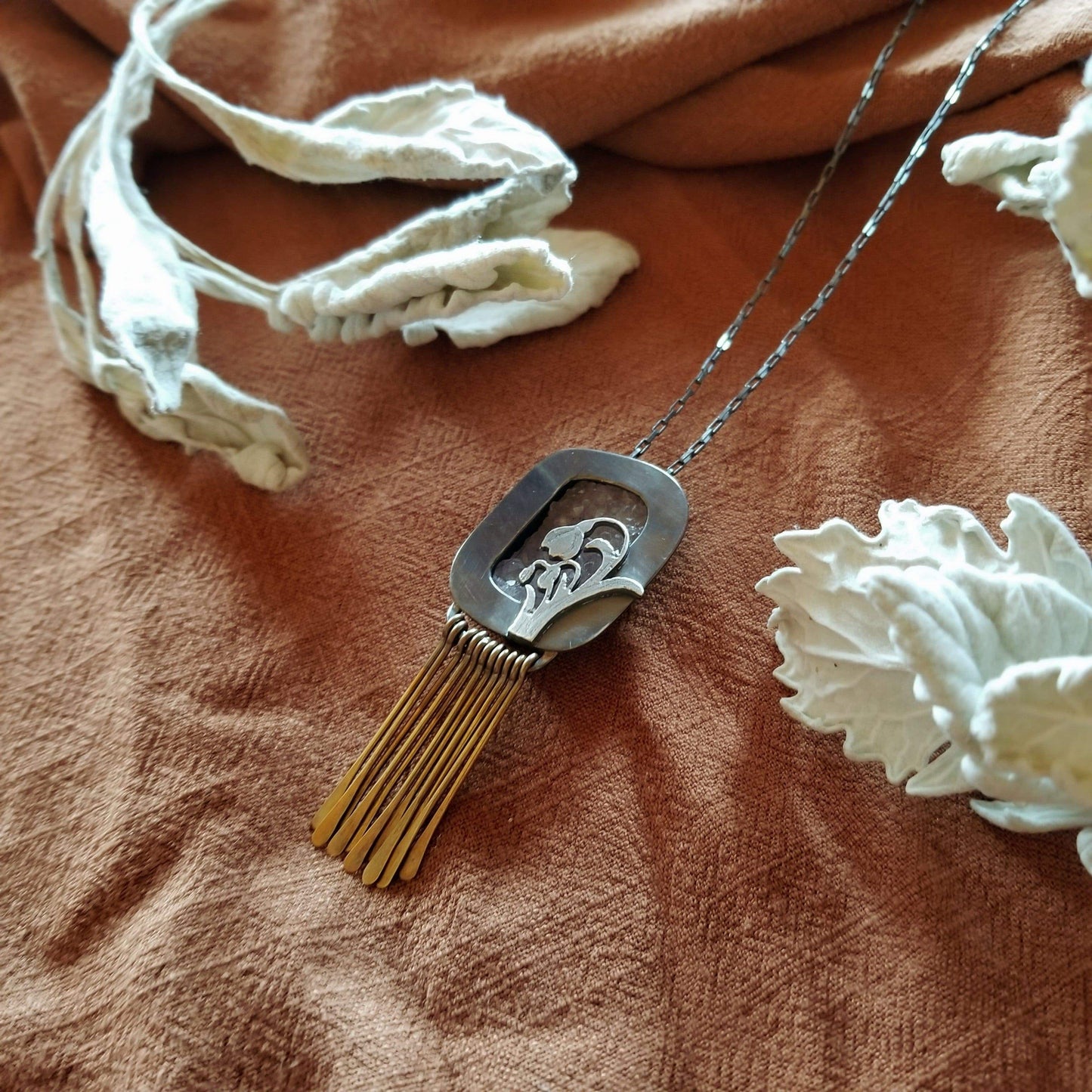 Right side angle - Snowdrop Necklace No. 3 - A rectangular amethyst druzy (pale purple) set in a sterling silver box frame. A hand pierced and hand engraved snowdrop is soldered on the bottm left. Handmade brass fringe hangs below the box frame. All hanging on a sterling silver chain.