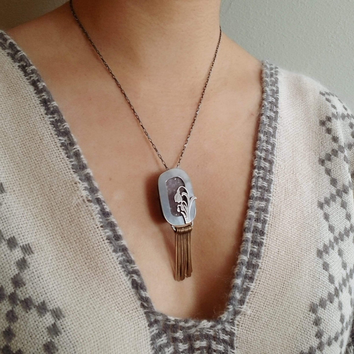 Model wearing Snowdrop Necklace No. 3 - A retangular amethyst druzy (pale purple) set in a sterling silver box frame. A hand pierced and hand engraved snowdrop is soldered on the bottm left. Handmade brass fringe hangs below the box frame. All hanging on a sterling silver chain.