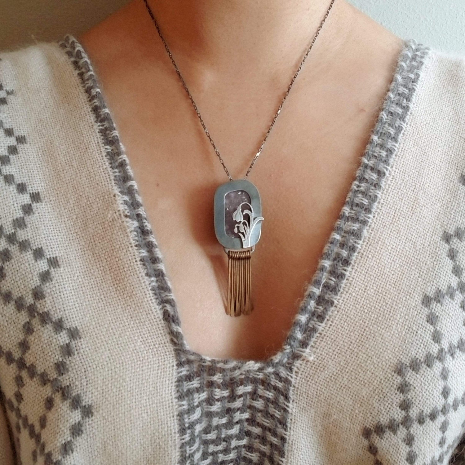 Model wearing Snowdrop Necklace No. 3 - A retangular amethyst druzy (pale purple) set in a sterling silver box frame. A hand pierced and hand engraved snowdrop is soldered on the bottm left. Handmade brass fringe hangs below the box frame. All hanging on a sterling silver chain.